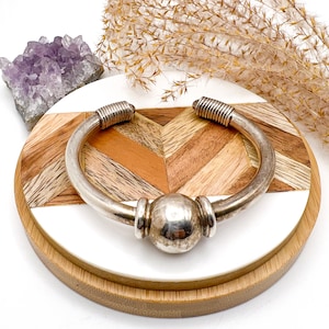 May include: A silver-toned cuff bracelet with a central sphere and ribbed details at the ends. The bracelet is displayed on a round wooden coaster with a chevron pattern of light and dark wood, and a white section.