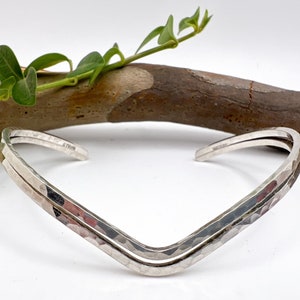 Può includere: Un bracciale a polsino argentato con una texture martellata e un design a forma di V. Il bracciale è aperto alle estremità e ha una costruzione a doppia fascia. Lo sfondo include un ramo di legno e foglie verdi.