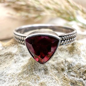May include: A silver ring with a triangular, deep red gemstone. The band has a textured design. The ring is set against a neutral, textured background, with soft, blurred greenery in the background.