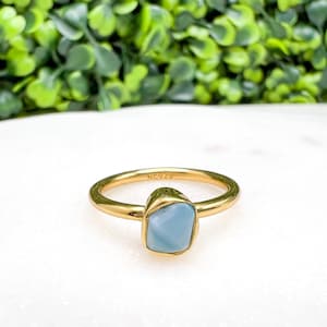 May include: A gold-colored ring with a light blue, rectangular stone. The ring is set on a white surface with a blurred green background. The ring is stamped with "NC925".