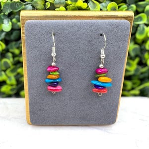 Könnte beinhalten: Ein Paar bunte Hängeohrringe. Jeder Ohrring hat einen Stapel lebendiger, oval geformter Perlen in den Farbtönen Rosa, Grün, Orange, Blau und Rot. Die Ohrringe hängen an silberfarbenen Haken und schaffen ein verspieltes und auffälliges Accessoire.