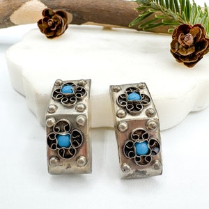 May include: A pair of silver-toned earrings with a rectangular shape. Each earring features a central turquoise-colored stone surrounded by a black floral design. The edges are adorned with small, round studs.