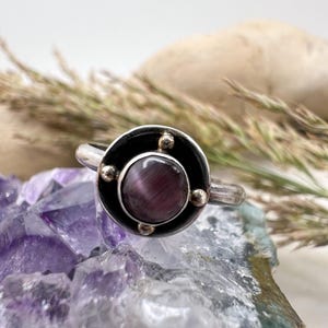 May include: A silver ring with a dark purple, round gemstone set in a black and silver circular design. The ring has small, gold-colored accents around the gemstone. The ring is displayed on a purple crystal surface.