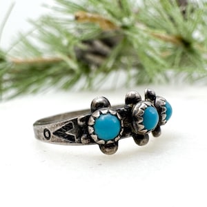 May include: A silver ring featuring three bright turquoise stones. The ring has a detailed, vintage design with decorative elements around the band and the turquoise settings. The ring is set against a white background with a blurred green branch.