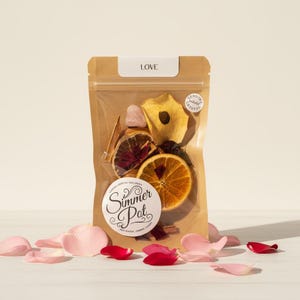 Può includere: Un sacchetto di carta marrone con un fronte trasparente, etichettato "LOVE", contiene fette d'arancia essiccate, pezzi di mela, bastoncini di cannella e un cristallo di quarzo rosa. Un adesivo recita "Simmer Pot". Petali di rosa rosa e rossi sono sparsi intorno al sacchetto.