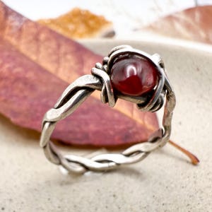 May include: A silver ring with a woven band and a round, deep red gemstone. The gemstone is held in place by a wire-wrapped setting. The ring is set against a blurred background of a leaf.