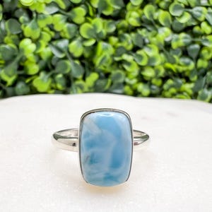 May include: A rectangular, light blue larimar gemstone ring with a silver band. The gemstone has a cloudy appearance with white swirls. The ring is set against a white surface with a green leafy background.