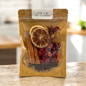 Może przedstawiać: Przezroczysta torebka wypełniona świątecznymi składnikami. Zawiera suszone plasterki pomarańczy, laski cynamonu, anyż gwiaździsty i żurawinę. Torebka jest zamknięta etykietą z napisem "HOME FOR THE HOLIDAYS".