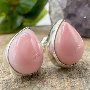 Könnte beinhalten: Ein Paar silberner Ohrringe mit tropfenförmigen, polierten rosa Edelsteinen. Die Ohrringe sind in Silber gefasst und haben eine glatte, glänzende Oberfläche. Die Ohrringe werden auf einer strukturierten Oberfläche mit grünem Laub im Hintergrund präsentiert.