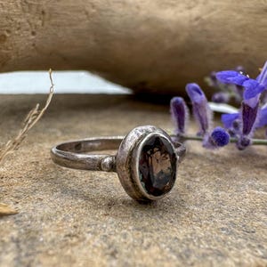 May include: A silver ring featuring an oval-shaped, dark brown faceted gemstone set in a simple metal bezel. The band has a weathered, oxidized finish with a small spherical detail where the band meets the setting, showcasing a classic design.