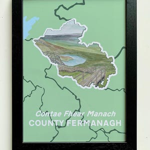 May include: A framed print of a map of County Fermanagh, Ireland. The map is green with a white outline. The county name is written in both English and Irish. The map is surrounded by a black frame.