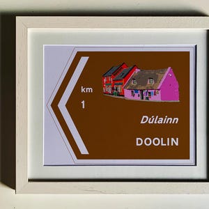 May include: A brown and white sign with a white arrow pointing to the right. The sign has the text "Dúlainn DOOLIN" and "1 km" on it. The sign also has a picture of a row of colorful houses.