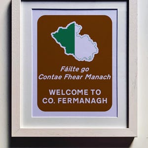 May include: A framed print with a brown background and a white outline. The print features a green and white map of County Fermanagh, Northern Ireland. The text "Fáilte go Contae Fhear Manach" is written in Irish Gaelic, and the text "Welcome to Co. Fermanagh" is written in English.