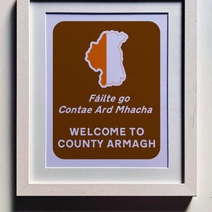 Puede incluir: Una impresión enmarcada con fondo marrón y contorno blanco. La impresión presenta un mapa del condado de Armagh, Irlanda del Norte, con el texto "Fáilte go Contae Ard Mhacha" en gaélico irlandés y "Welcome to County Armagh" en inglés.