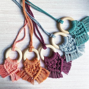 Puede incluir: Una colección de juguetes de gimnasio para bebés hechos a mano con macramé en varios colores, incluyendo coral, naranja, morado, turquesa y gris. Cada juguete presenta un anillo de madera y un diseño de macramé anudado con flecos. Los juguetes están suspendidos por cuerdas a juego.