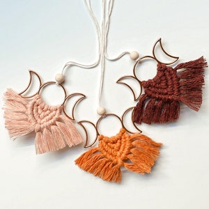 Puede incluir: Tres tapices de pared de macramé con fases lunares con cuentas de madera y aros de metal dorado. Los tapices son en tonos melocotón, naranja y marrón.