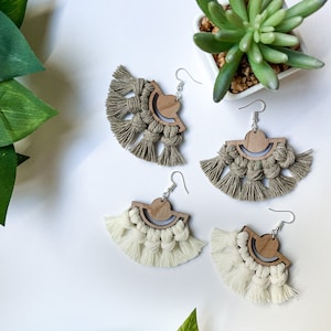 May include: Four pairs of handmade macrame earrings. Each earring features a wooden semi-circle design with a knotted fringe. Two pairs are beige, and two are a muted olive green. The earrings are displayed on a white surface.