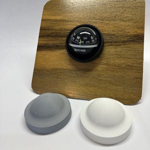 Puede incluir: Una brújula negra con una cara blanca y la palabra "RITCHIE" impresa en ella. La brújula está montada sobre una base de madera. Hay dos objetos redondos, grises y blancos, impresos en 3D en primer plano.