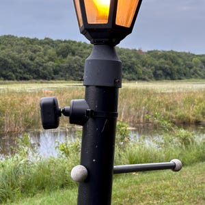 May include: A black metal lamp post featuring a glowing amber light fixture at the top, a small black camera mounted to the side with a ball joint, and a horizontal metal bar with two light gray spherical ends attached to the main pole.