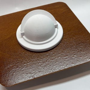 May include: A white, 3D-printed object with a rounded top and a circular base. The object is set on a square, wooden surface with a glossy, brown finish. The object has a smooth texture.