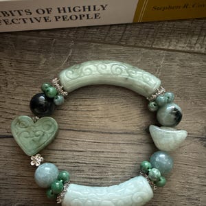 May include: A jade bracelet featuring a heart-shaped charm, carved cylindrical beads, and round beads in shades of green and blue. The bracelet is accented with silver-colored metal and rests on a wooden surface.