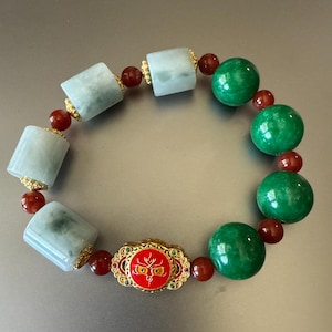 May include: A beaded bracelet featuring alternating light blue, green, and red beads. The bracelet includes cylindrical and spherical beads, with a central gold-colored charm with a red face design. The bracelet is a piece of jewelry.
