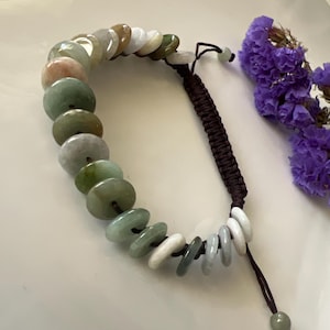 May include: A jade bracelet with a brown cord and a variety of green, white, and pink jade beads. The beads are round and flat, and they are strung on a cord that is tied with a knot at the end.