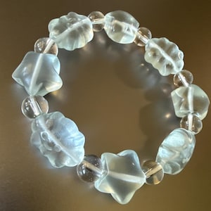 May include: A bracelet featuring clear, light blue, and clear beads. The bracelet includes star and flower-shaped beads, interspersed with round, clear beads. The bracelet is displayed on a reflective surface.