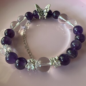 May include: A purple and clear crystal bracelet with a silver butterfly charm. The bracelet is made with amethyst and clear quartz beads.
