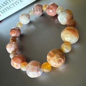 May include: A beaded bracelet featuring a mix of faceted and round beads in shades of orange, peach, and cream. The bracelet is made of natural stones and is approximately 7 inches in circumference. The book in the background reads "The 7 Habits of Effective People."