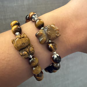 May include: Two bracelets with tiger's eye beads and a carved cat charm. The bracelets are brown and gold with silver accents.