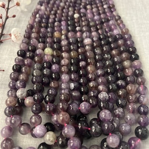 May include: A strand of purple amethyst beads, each bead is round and smooth. The beads are strung on a thin thread and are arranged in a pile.