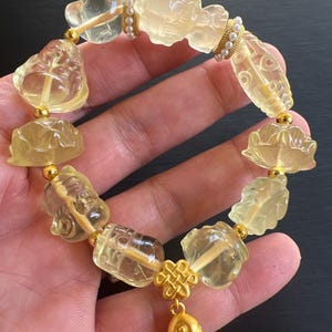 May include: A yellow crystal bracelet with gold accents and a gold fish charm. The bracelet features carved crystal beads in the shape of a lotus flower, a Buddha, and other lucky symbols.