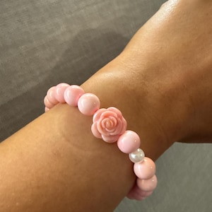 May include: Pink beaded bracelet featuring a central rose-shaped bead and interspersed round beads. The bracelet includes a single white pearl bead. The beads are strung together on an elastic band, creating a delicate and feminine accessory.