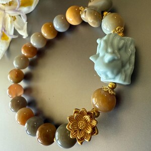 May include: A jade bracelet with a light blue jade charm and a gold flower charm. The bracelet is made of round beads in shades of brown, gray, and beige.