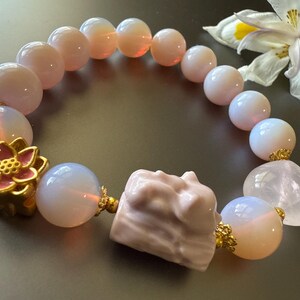 May include: A pink and white gemstone bracelet with a gold flower charm and a pink and white gemstone charm. The bracelet is made of round beads and has a delicate, feminine look.