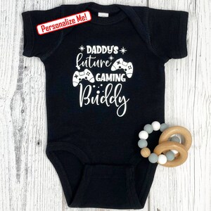 May include: Black baby onesie with white text that reads "*Daddy's future* gaming buddy" with a graphic of two video game controllers.