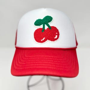 May include: A red and white trucker hat with a cherry patch. The hat features a red brim and a white front panel with a textured red cherry design and green stem. The hat has a red rope detail.