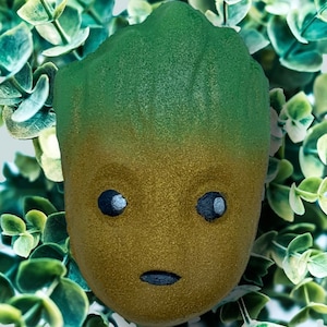 May include: A Groot-shaped bath bomb, with a green top and golden-brown base. The eyes are black with white accents, and the mouth is a black line. The bath bomb is set against a backdrop of green foliage, creating a natural aesthetic.