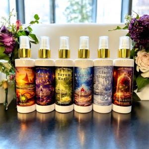 May include: Six white spray bottles with gold tops and fairytale-themed labels. The labels feature text including "Wayfinder", "Arabah Nights", and "Enchantée". The bottles are displayed in a row, with floral arrangements on either side.