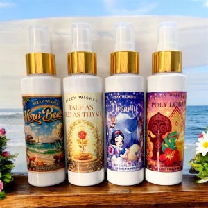 May include: Four white spray bottles with gold caps, each labelled "Fizzy Wishes." The labels feature illustrations and text: "Vero Beach," "Tale as Old as Thyme," "Dreamy," and "Poly Lobby." The background shows a beach scene.