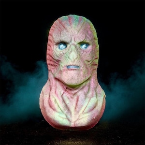 May include: A monster head-shaped bath bomb with a textured surface. The bath bomb is coloured pink, green, and blue, with glowing blue eyes and a blue mouth. The background is dark with a hint of blue mist, creating a spooky atmosphere.