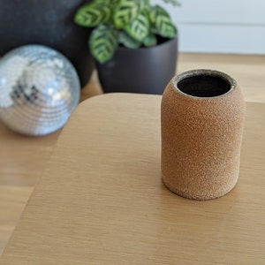 May include: A small, brown, textured ceramic vase with a black interior. The vase is sitting on a light brown wooden surface.