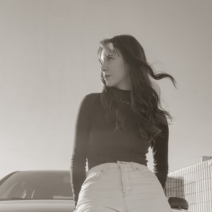 Peut inclure: Une femme aux longs cheveux bruns portant un pull noir &agrave; col roul&eacute; et un jean bleu clair. Elle se tient devant une voiture et regarde sur le c&ocirc;t&eacute;.