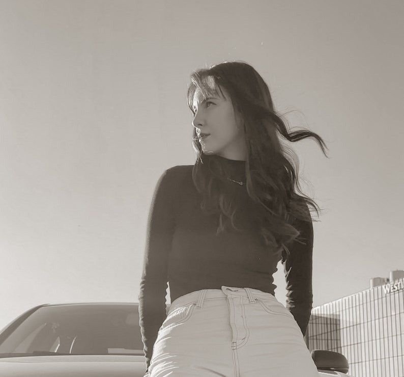 Peut inclure: Une femme aux longs cheveux bruns portant un pull noir &agrave; col roul&eacute; et un jean bleu clair. Elle se tient devant une voiture et regarde sur le c&ocirc;t&eacute;.