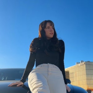 Peut inclure: Une femme portant un chemisier noir &agrave; col roul&eacute; et un jean beige clair est pench&eacute;e sur le capot d'une voiture noire. Le ciel est bleu et il y a un b&acirc;timent en arri&egrave;re-plan.