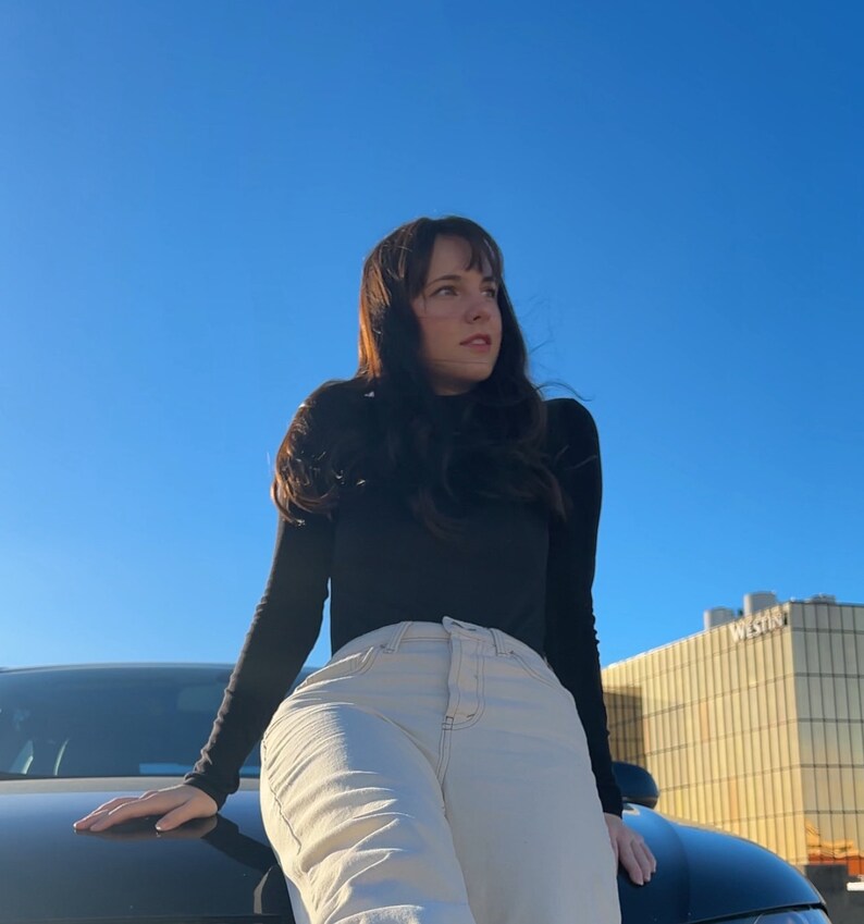Peut inclure: Une femme portant un chemisier noir &agrave; col roul&eacute; et un jean beige clair est pench&eacute;e sur le capot d'une voiture noire. Le ciel est bleu et il y a un b&acirc;timent en arri&egrave;re-plan.