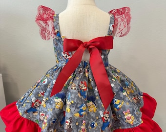 May include: A gray dress featuring a pattern of Mario characters, with red accents. The dress has a red bow in the back and red ruffles at the hem. The sleeves are adorned with red lace.