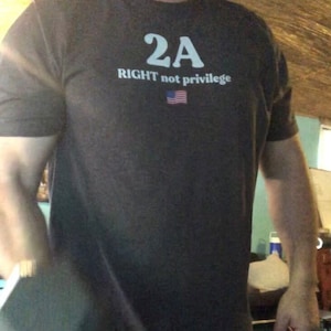 Può includere: Una maglietta grigia scura con il testo "2A RIGHT not privilege" e una piccola grafica della bandiera americana.