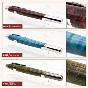May include: Three wooden pens with different colored wood grain patterns. The pens are on a white surface with a clear stand. The pens are labeled R-001 DD Pink/Blue Burl, R-004 Blue Curly Maple, and R-005 DD Green Maple Burl.